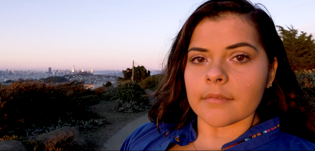 A young woman looking at the camera, with the sunset behind her.